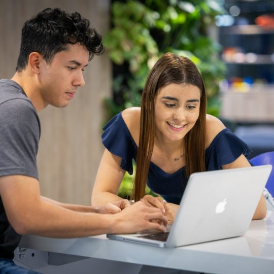 Dos personas están sentadas frente a una mesa utilizando una computadora portátil plateada, aparentemente colaborando o revisando información en la pantalla.