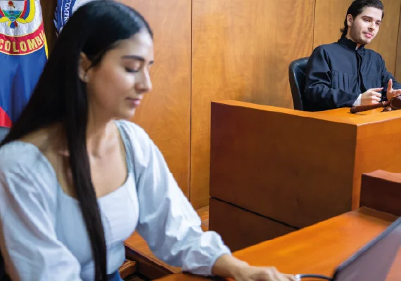 Escenario de simulación judicial con una persona escribiendo en un computador portátil en primer plano y otra persona en el estrado, detrás de un escritorio, con banderas oficiales al fondo.