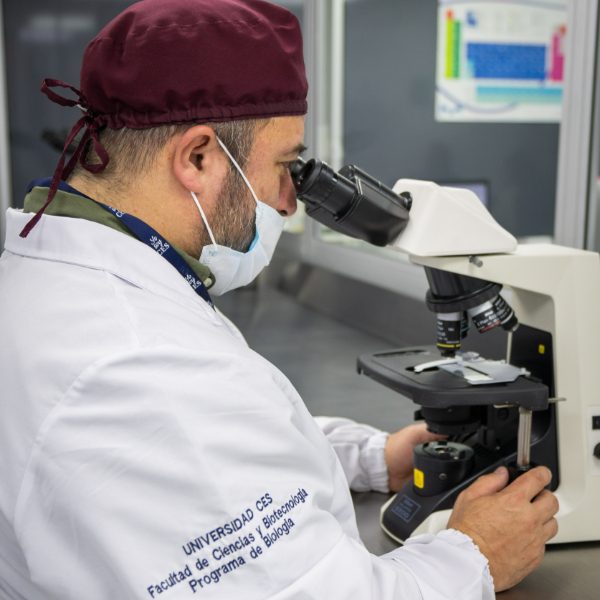 Persona con bata blanca y gorro oscuro utiliza un microscopio en un laboratorio, sobre una mesa con equipo científico y documentos visibles.