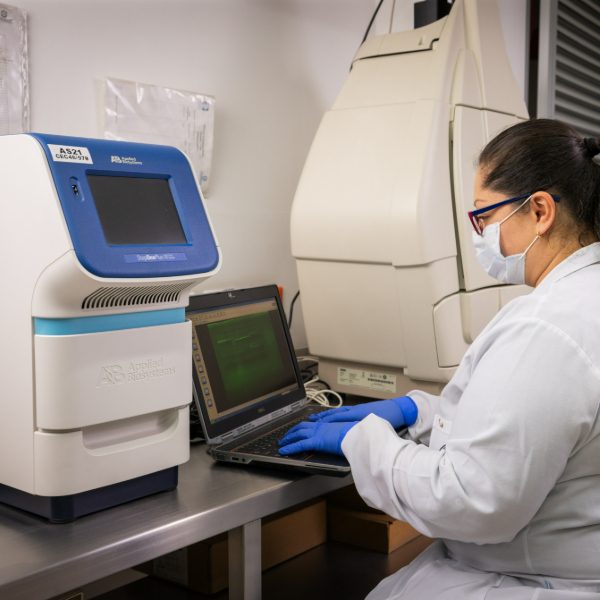 Persona con bata blanca y guantes azules trabaja en un laboratorio, utilizando un computador portátil junto a un equipo especializado de análisis biomolecular.