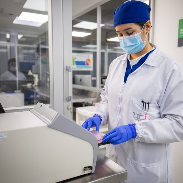 Persona con bata de laboratorio y guantes azules operando un equipo especializado en un entorno limpio y controlado.