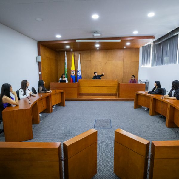 Sala de audiencias simulada con estrados de madera dispuestos en forma de U. En el centro, una persona en el estrado principal señala hacia un lado, mientras otras personas están sentadas en los estrados laterales. Al fondo se observan banderas oficiales y paredes de madera.