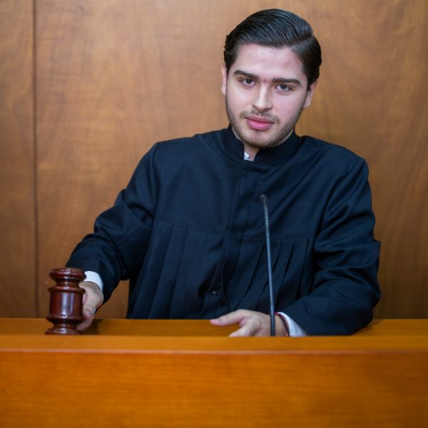 Persona con toga judicial está de pie en un estrado de madera, sosteniendo un mazo junto a un micrófono, en un entorno que simula una sala de audiencias.