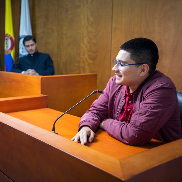 Persona con chaqueta roja está sentada en un estrado frente a un micrófono, en una sala que simula un juicio, con banderas oficiales y mobiliario de madera al fondo.
