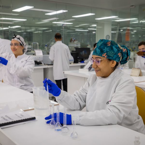 Personas en laboratorio realizan prácticas con material de vidrio y líquidos, usando batas blancas, guantes azules y gorros de protección, en un espacio con mesas y equipos especializados.