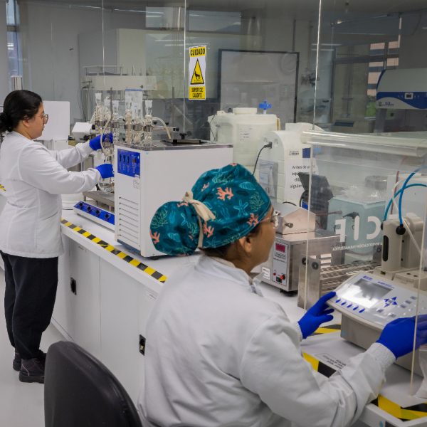 Dos personas en laboratorio operan equipos de análisis con frascos y tubos conectados, usando batas blancas, guantes azules y gorros de protección, en un entorno con superficies limpias y señal de advertencia visible.