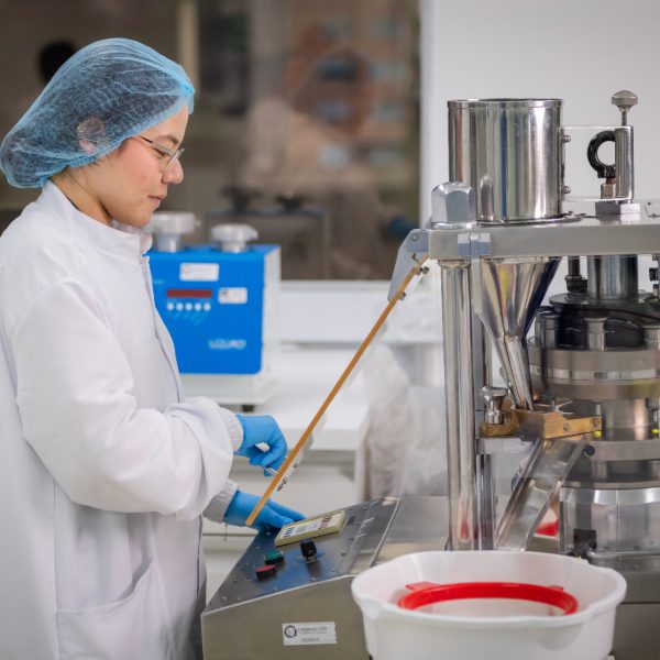 Persona en laboratorio opera una máquina industrial para procesamiento de alimentos, con recipientes y líquidos visibles, usando bata blanca, gorro y guantes azules.