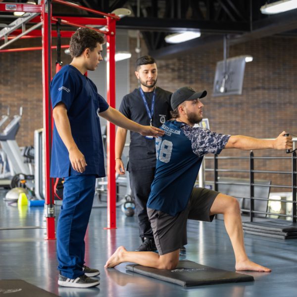 Una persona realiza un ejercicio de fuerza y estabilidad en posición de rodillas sobre una colchoneta, sosteniendo una cuerda o barra, mientras dos personas con uniforme clínico observan y asisten en un gimnasio con equipos de entrenamiento al fondo.