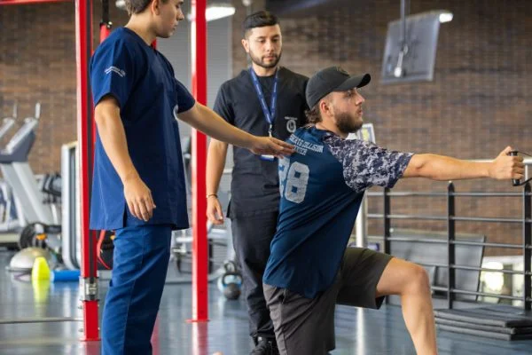 Una persona realiza un ejercicio de fuerza y estabilidad en posición de rodillas sobre una colchoneta, sosteniendo una cuerda o barra, mientras dos personas con uniforme clínico observan y asisten en un gimnasio con equipos de entrenamiento al fondo.