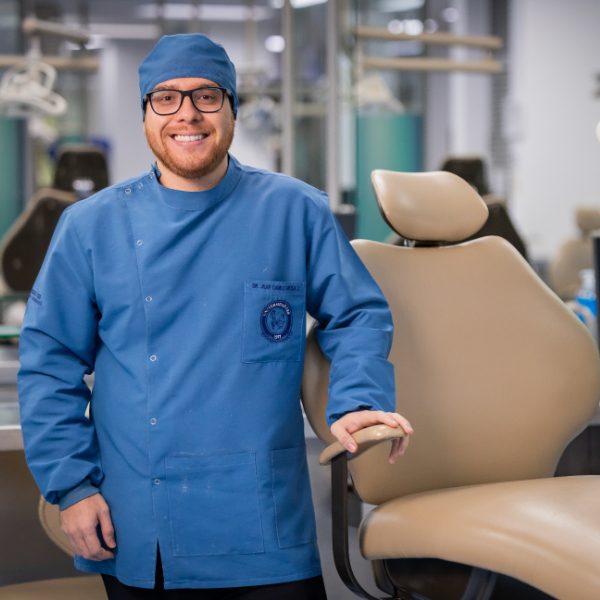 Una persona con uniforme odontológico azul está de pie junto a una silla dental en un consultorio equipado para prácticas clínicas.