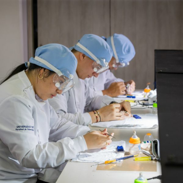 Varias personas con batas blancas y gorros azules trabajan en una mesa realizando prácticas manuales con instrumentos odontológicos y materiales, en un entorno académico o laboratorio.