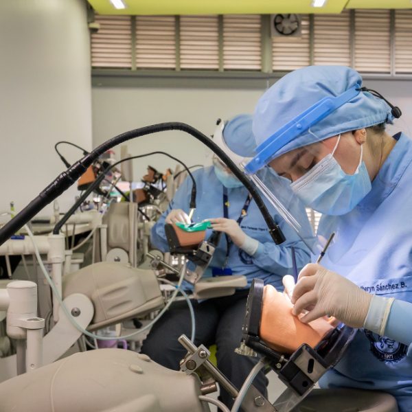 Personas con uniforme clínico azul y gorros realizan prácticas odontológicas en simuladores, utilizando instrumentos dentales en un aula equipada.