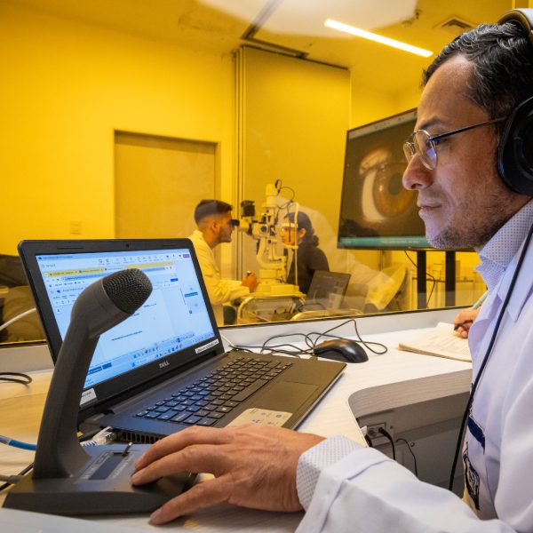 Docente observa imágenes clínicas en un computador mientras guía un examen ocular realizado por dos profesionales en un consultorio separado por vidrio.