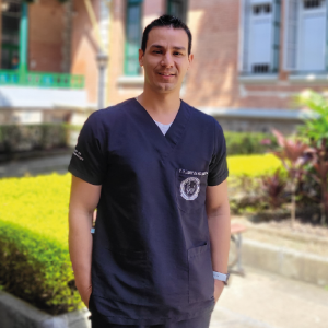 Persona de pie en un jardín con vegetación y edificio al fondo, usando uniforme médico de color azul oscuro con logotipo bordado en el pecho y reloj en la muñeca izquierda.