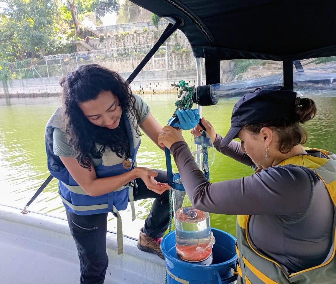 Dos personas en una embarcación recolectando muestras de agua con equipo especializado, en un cuerpo de agua rodeado de vegetación y estructuras.