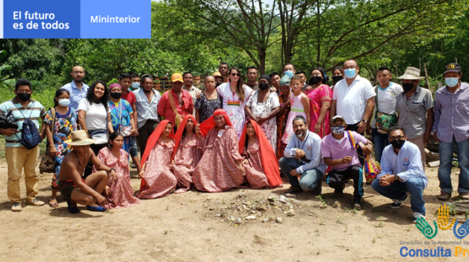 Grupo numeroso de personas posando al aire libre en un entorno natural, algunas con vestimenta tradicional colorida y otras con ropa casual, junto a carteles institucionales en la imagen.