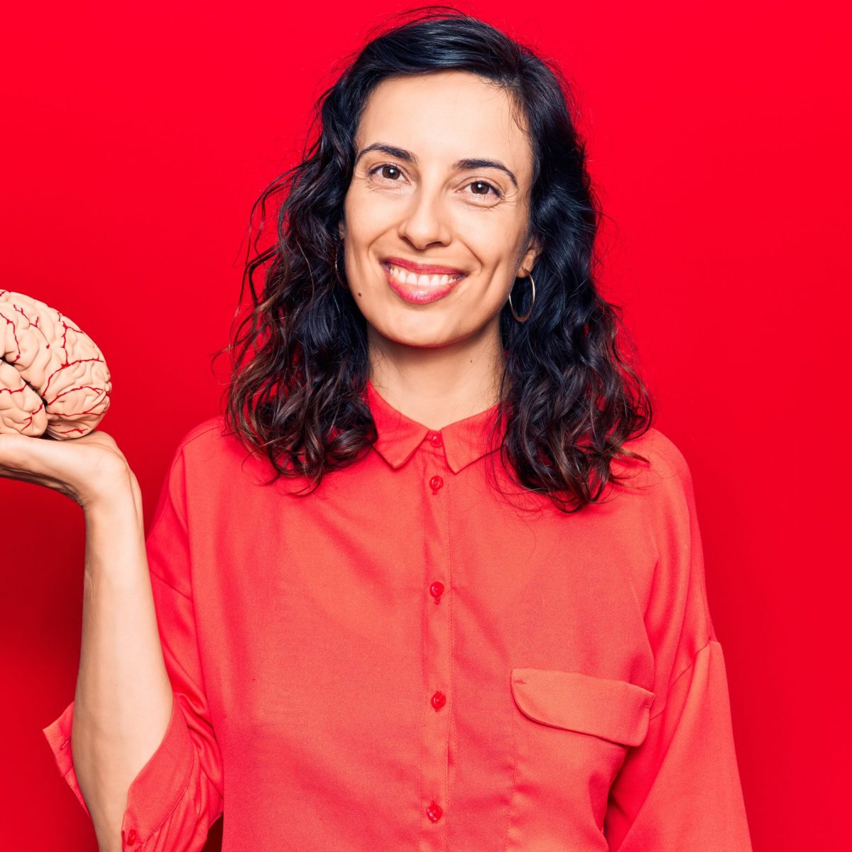Persona vestida con camisa roja sostiene un modelo anatómico de un cerebro en una mano, sobre fondo rojo.