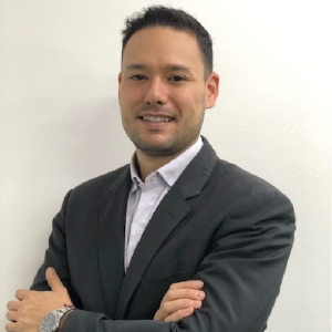 La imagen muestra a una persona de pie frente a una pared blanca, con los brazos cruzados. Viste un saco formal oscuro y una camisa clara abotonada. También lleva un reloj metálico en la muñeca izquierda.