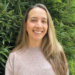 La imagen muestra a una persona de pie frente a un fondo de hojas verdes. Lleva un suéter de color beige claro con textura suave y el cabello largo, lacio y castaño con reflejos claros, peinado hacia un lado.