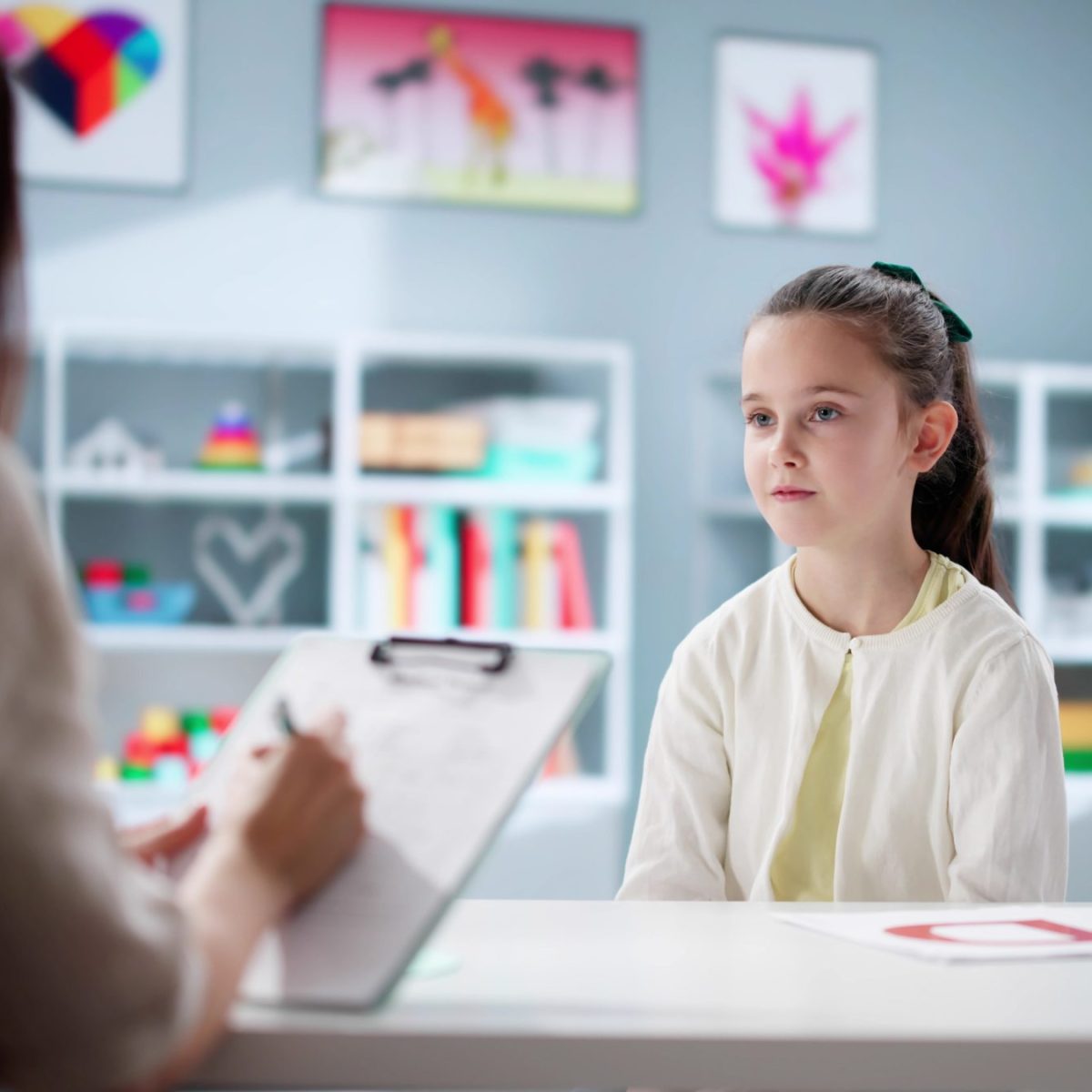 En un consultorio, una persona adulta sostiene una carpeta mientras conversa con una niña sentada frente a ella; sobre la mesa hay una hoja con una letra grande y al fondo se observan estanterías con libros, juguetes y decoraciones coloridas.