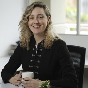 La imagen muestra a una persona sentada en una oficina, sosteniendo una taza blanca con ambas manos sobre un escritorio. Viste una chaqueta negra con botones metálicos decorativos en dos filas verticales. En el escritorio hay una pequeña planta en maceta blanca y el fondo incluye una ventana que deja entrar luz natural.