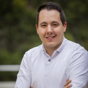 La imagen muestra a una persona con los brazos cruzados, vistiendo una camisa blanca de manga larga con cuello tipo mao y botones visibles. En la muñeca izquierda lleva un reloj inteligente con correa azul. El fondo es exterior, con vegetación desenfocada en tonos verdes.