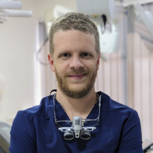 La imagen muestra a una persona de pie en un consultorio odontológico, vistiendo uniforme clínico azul oscuro. Lleva un equipo de aumento con luz frontal colgado al cuello. El fondo incluye equipamiento dental y mobiliario médico en tonos claros, con iluminación blanca y ambiente clínico.