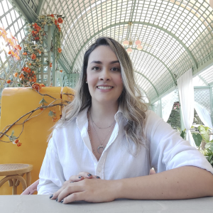 La imagen muestra a una persona sentada a una mesa en un espacio interior luminoso, con las manos apoyadas sobre la superficie. Viste una camisa blanca de manga larga y lleva collares finos. El entorno presenta una estructura metálica arqueada con techo traslúcido, decoración floral en tonos cálidos, plantas alrededor y cortinas blancas, lo que sugiere un ambiente tipo invernadero o café elegante con abundante luz natural.