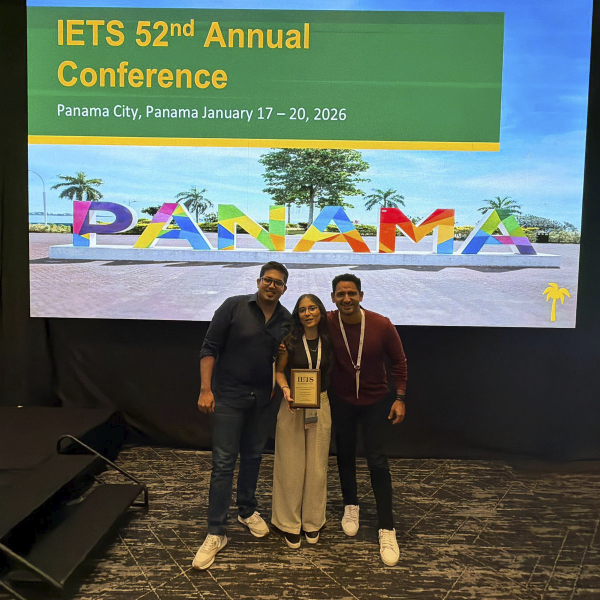 La imagen muestra a tres personas de pie en un escenario interior, posando juntas frente a una gran pantalla. En la pantalla se lee “IETS 52nd Annual Conference” y “Panama City, Panama January 17–20, 2026”, junto a una imagen colorida de la palabra “PANAMA”. Una de las personas sostiene un reconocimiento enmarcado. El entorno corresponde a un evento académico o institucional.