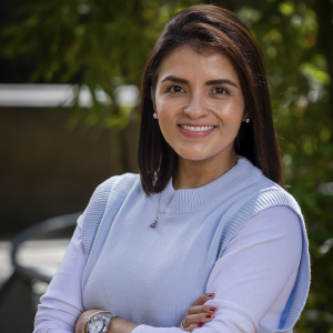 Persona de pie con los brazos cruzados, vistiendo un suéter claro con chaleco tejido azul y accesorios plateados, en un entorno exterior con vegetación.