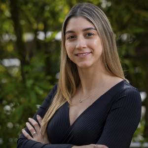 Persona de pie con los brazos cruzados, vestida con una blusa negra de escote en ‘V’ y joyería plateada, en un entorno exterior con vegetación.