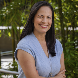 Persona de pie con un chaleco tejido azul claro y un collar largo plateado, en un entorno exterior con vegetación.