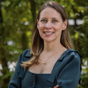 Persona de pie con los brazos cruzados, vistiendo un atuendo verde oscuro con mangas cortas y un collar dorado, en un entorno exterior con vegetación.
