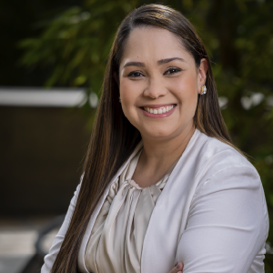 Persona de pie con los brazos cruzados, vistiendo una chaqueta y blusa en tonos claros, con accesorios plateados, en un entorno exterior con vegetación.