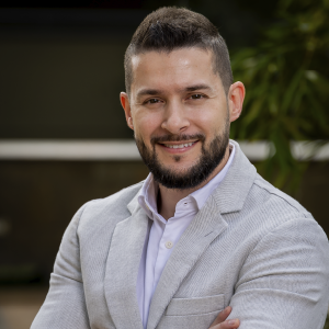 Persona de pie con los brazos cruzados, vistiendo una chaqueta gris claro sobre una camisa blanca, en un entorno exterior con vegetación.