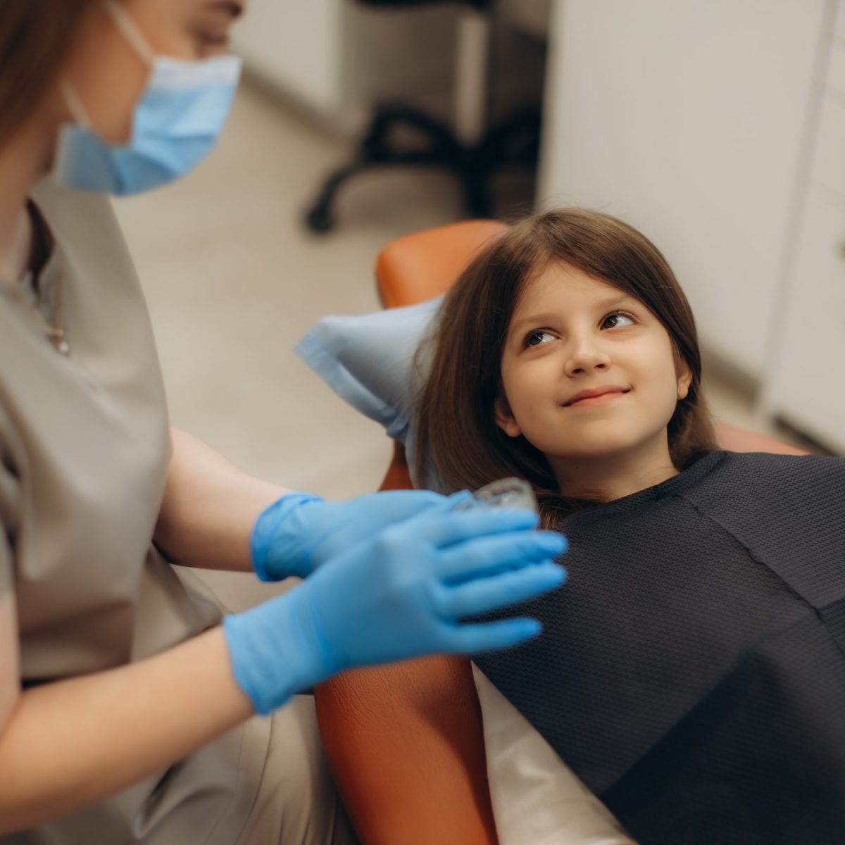 Una profesional de la salud dental atiende a una niña que se encuentra recostada en una silla odontológica, usando guantes y preparando instrumentos en un consultorio clínico.