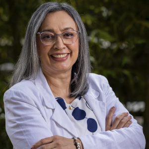 Persona de pie con los brazos cruzados, vistiendo una chaqueta clara sobre una blusa estampada en tonos azul y blanco, en un entorno exterior con vegetación.