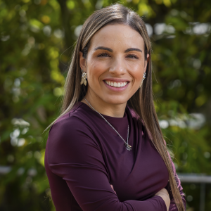 Persona de pie con los brazos cruzados, vistiendo una blusa color vino tinto y accesorios plateados, en un entorno exterior con vegetación.