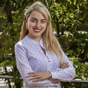 Persona de pie con los brazos cruzados, vistiendo una blusa clara con estampado de puntos y accesorios dorados, en un entorno exterior con vegetación.