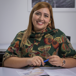 Persona sentada frente a una mesa, con un chal verde de mariposas y un reloj dorado, sosteniendo un bolígrafo sobre un documento abierto.