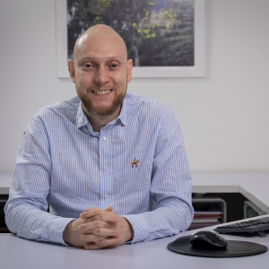Persona sentada frente a un escritorio, vistiendo una camisa de rayas azules y blancas, con las manos entrelazadas y un computador a un costado.