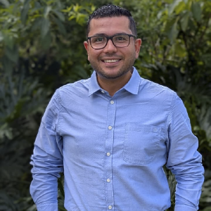 Persona de pie vistiendo una camisa azul claro de manga larga y botones, en un entorno exterior con vegetación.