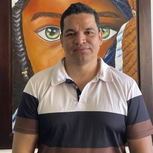 Persona de pie vistiendo una camiseta tipo polo con franjas en tonos blanco, negro y marrón, frente a un cuadro colorido con un ojo grande como elemento central.