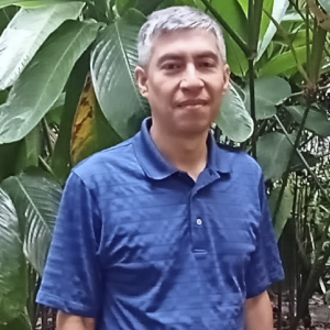 Persona de pie vistiendo una camiseta tipo polo azul, en un entorno exterior con abundante vegetación.