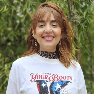 Persona de pie vistiendo una camiseta blanca con un estampado que incluye texto y colores llamativos, junto a un cuello alto de estampado animal, en un entorno exterior con vegetación.