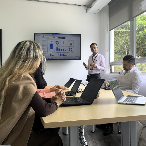La imagen muestra a un grupo de personas reunidas en una sala de reuniones, sentadas alrededor de una mesa con computadores portátiles abiertos. Una persona de pie realiza una presentación utilizando una pantalla con gráficos y diagramas. La sala tiene iluminación natural proveniente de una ventana lateral con vista a vegetación exterior.