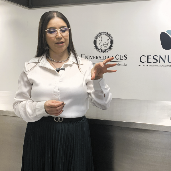 La imagen muestra a una persona de pie frente a un panel institucional con los logos de la Universidad CES y CESNUTRAL. Lleva una blusa blanca y una falda plisada oscura, y realiza un gesto con la mano mientras sostiene un objeto pequeño en la otra. El entorno corresponde a un espacio formal o académico.