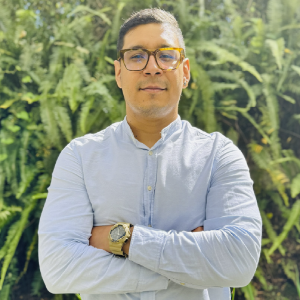 La imagen muestra a una persona de pie en un entorno exterior con abundante vegetación verde al fondo. Viste una camisa clara de manga larga y mantiene los brazos cruzados. Lleva un reloj metálico en la muñeca izquierda. La iluminación es natural y brillante.