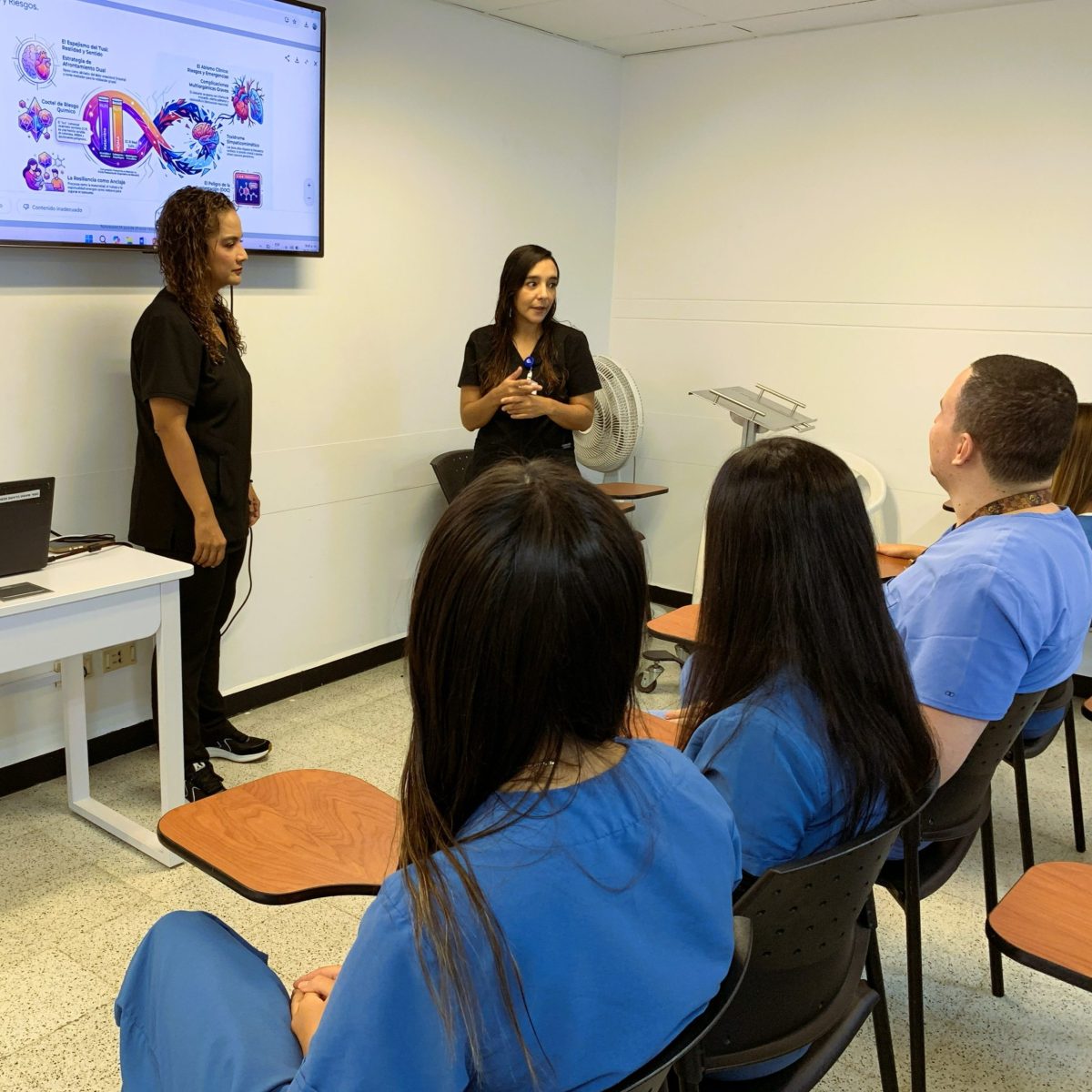 Un grupo de estudiantes en un aula observa una presentación proyectada en una pantalla, mientras dos personas al frente explican un tema relacionado con sustancias psicoactivas.