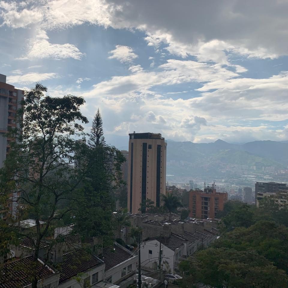 Vista urbana de la ciudad de Medellín con árboles en primer plano, varios edificios residenciales y una torre alta al centro, con montañas y cielo parcialmente nublado al fondo.